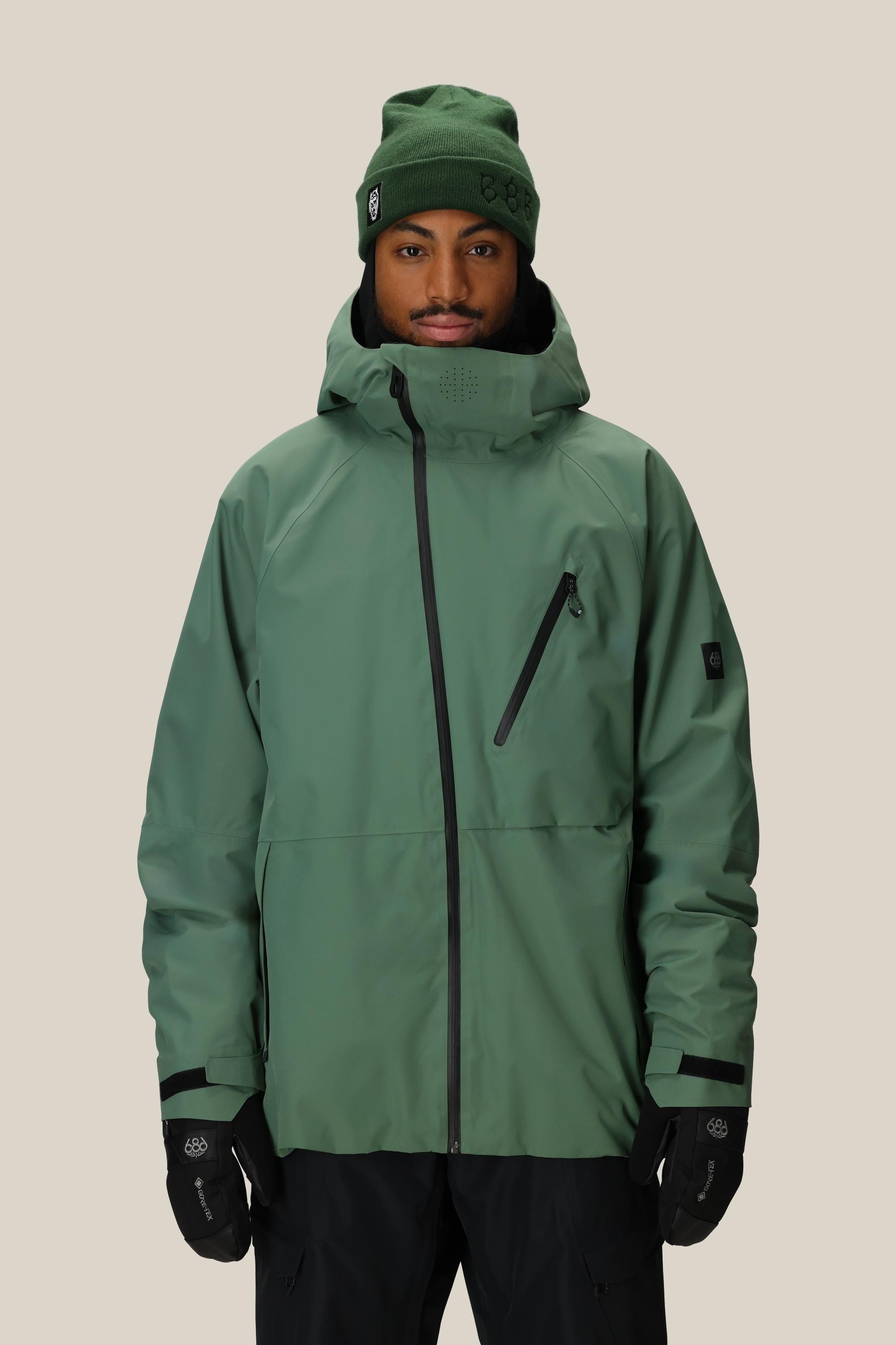 A model wears the 686 Men's Hydra™ Thermagraph® Jacket by 686 in green, paired with a green beanie and black gloves, standing against a beige background. The jacket features body-mapped lining, a zippered chest pocket, and black zipper details.