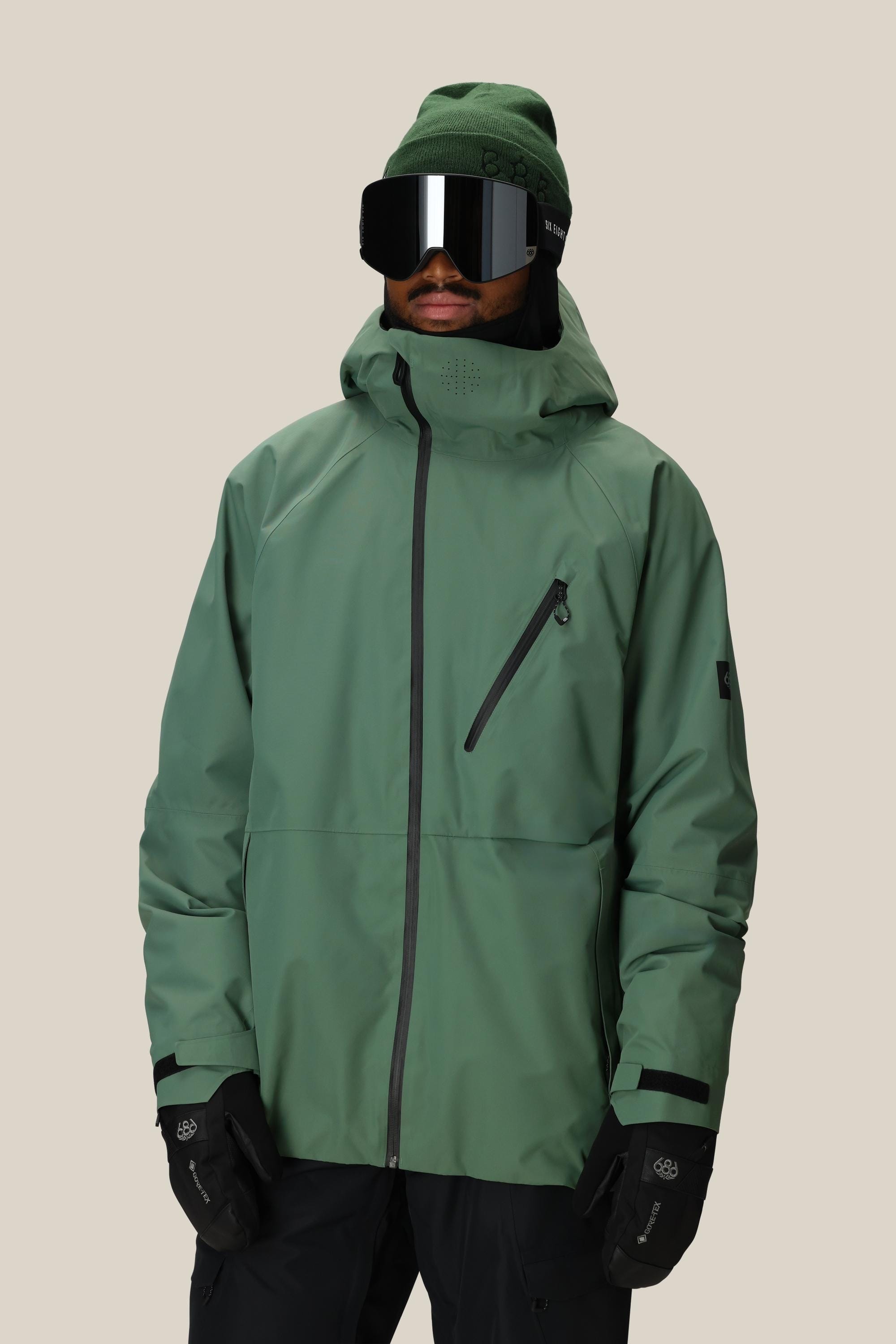 A person wears the 686 Men's Hydra™ Thermagraph® Jacket, a green beanie, black ski goggles, and black gloves from 686, standing against a plain beige background.
