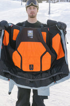 Wearing snow gear, a person stands on a snowy slope, displaying the 686 Men's Hydra™ Thermagraph® Jacket by 686 with its bright orange and black body-mapped lining for insulation. Ski goggles rest on their forehead.