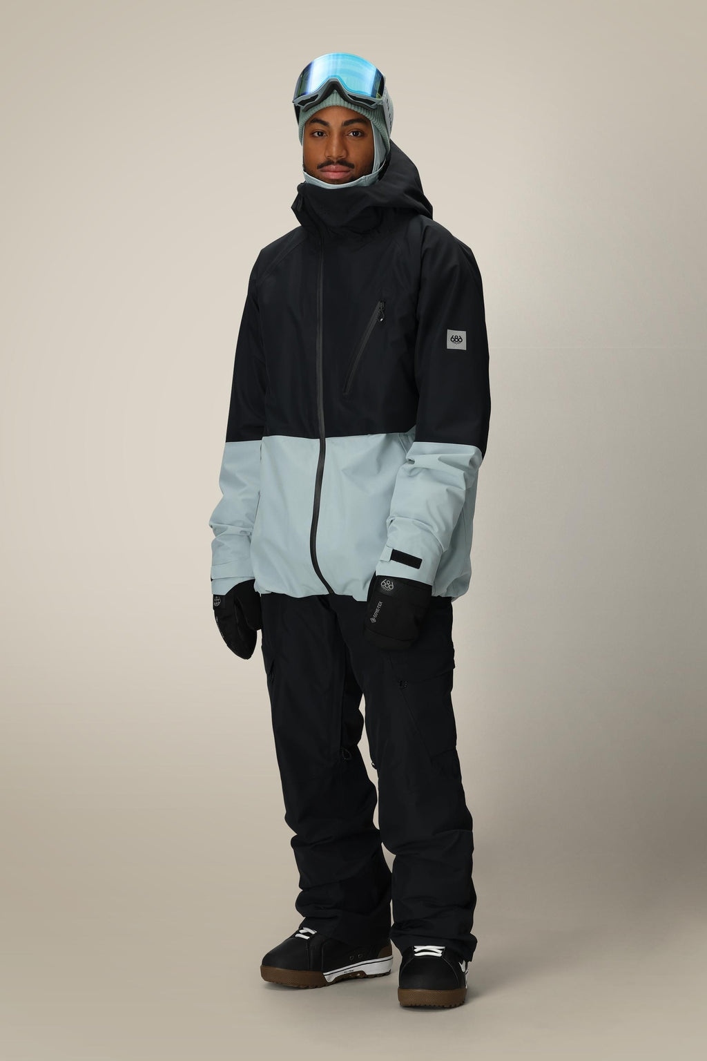 A person wearing the 686 Men's Hydra™ Thermagraph® Jacket with black snow pants, gloves, boots, and ski goggles stands against a plain beige background, facing forward with a neutral expression.