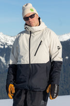 A person in the 686 Men's Hydra™ Thermagraph® Jacket, a best seller by 686 with body-mapped lining, stands outdoors in front of snow-covered mountains and a blue sky, wearing a beige beanie, dark sunglasses, and yellow gloves.