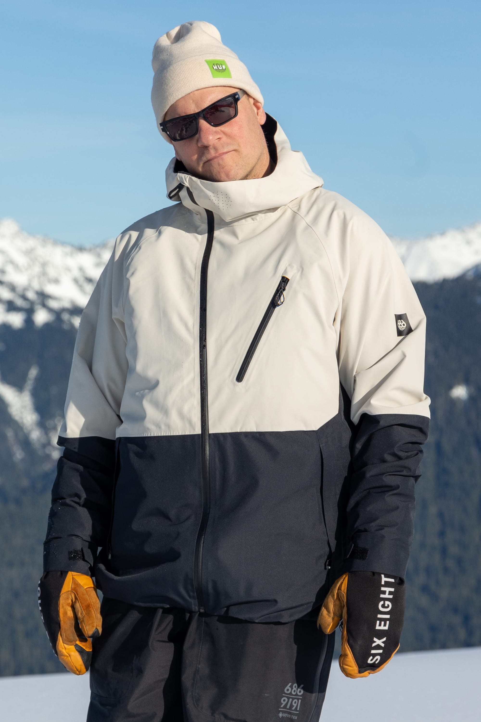A person in the 686 Men's Hydra™ Thermagraph® Jacket, a best seller by 686 with body-mapped lining, stands outdoors in front of snow-covered mountains and a blue sky, wearing a beige beanie, dark sunglasses, and yellow gloves.