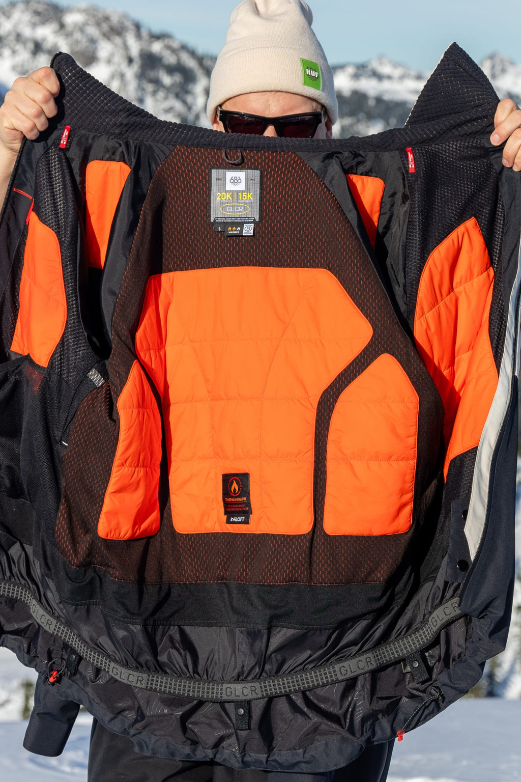 A person in sunglasses and a white beanie holds open the black and orange 686 Men's Hydra™ Thermagraph® Jacket, revealing its body-mapped lining and technical labels, with snowy mountains and blue sky in the background.