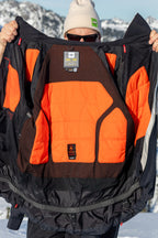 A person in sunglasses and a white beanie holds open the black and orange 686 Men's Hydra™ Thermagraph® Jacket, revealing its body-mapped lining and technical labels, with snowy mountains and blue sky in the background.