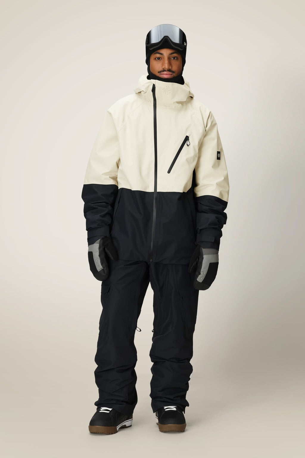 A person in the 686 Men's Hydra™ Thermagraph® Jacket by 686, paired with black snow pants, gloves, boots, and ski goggles, stands against a plain light background.