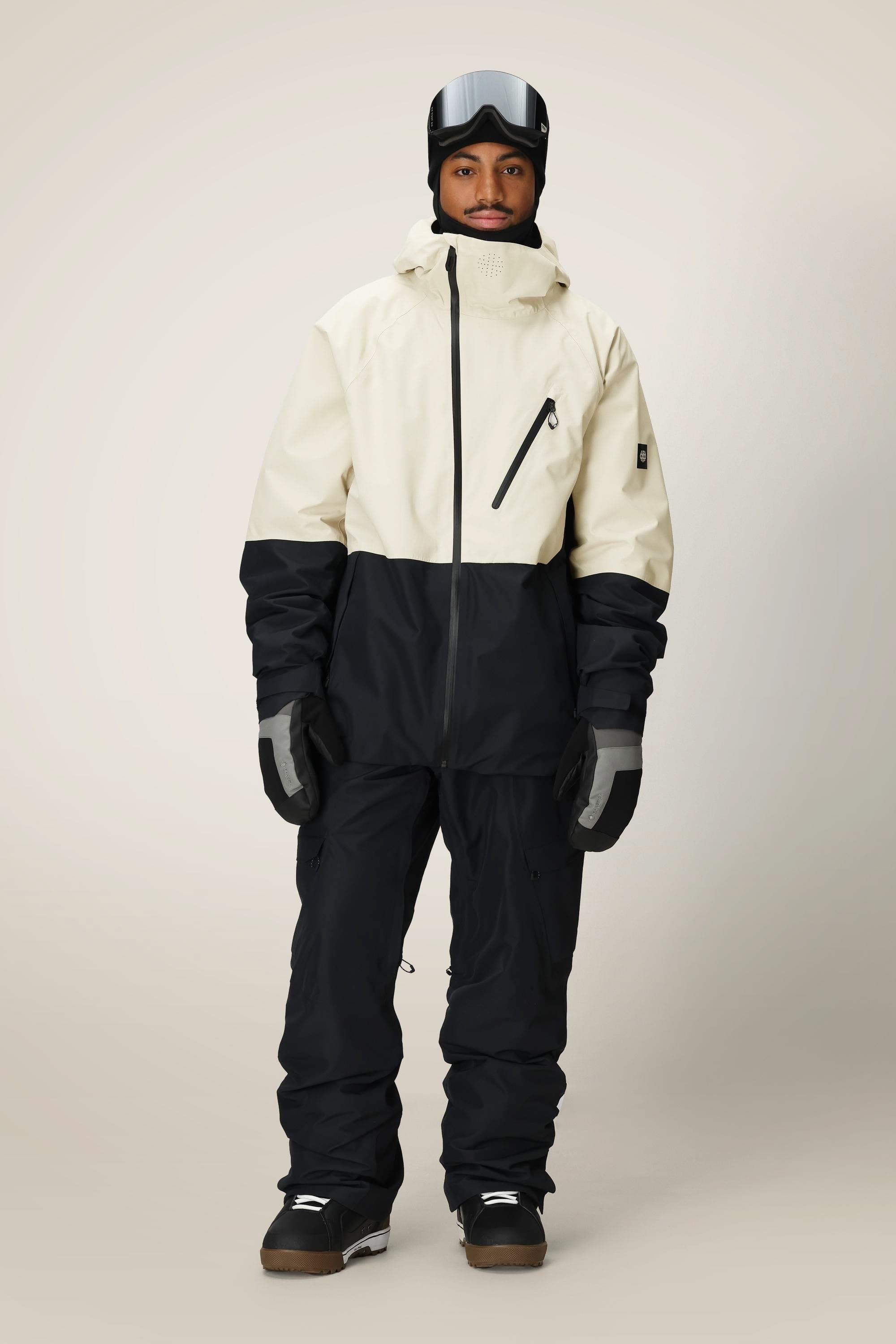 A person in the 686 Men's Hydra™ Thermagraph® Jacket by 686, paired with black snow pants, gloves, boots, and ski goggles, stands against a plain light background.