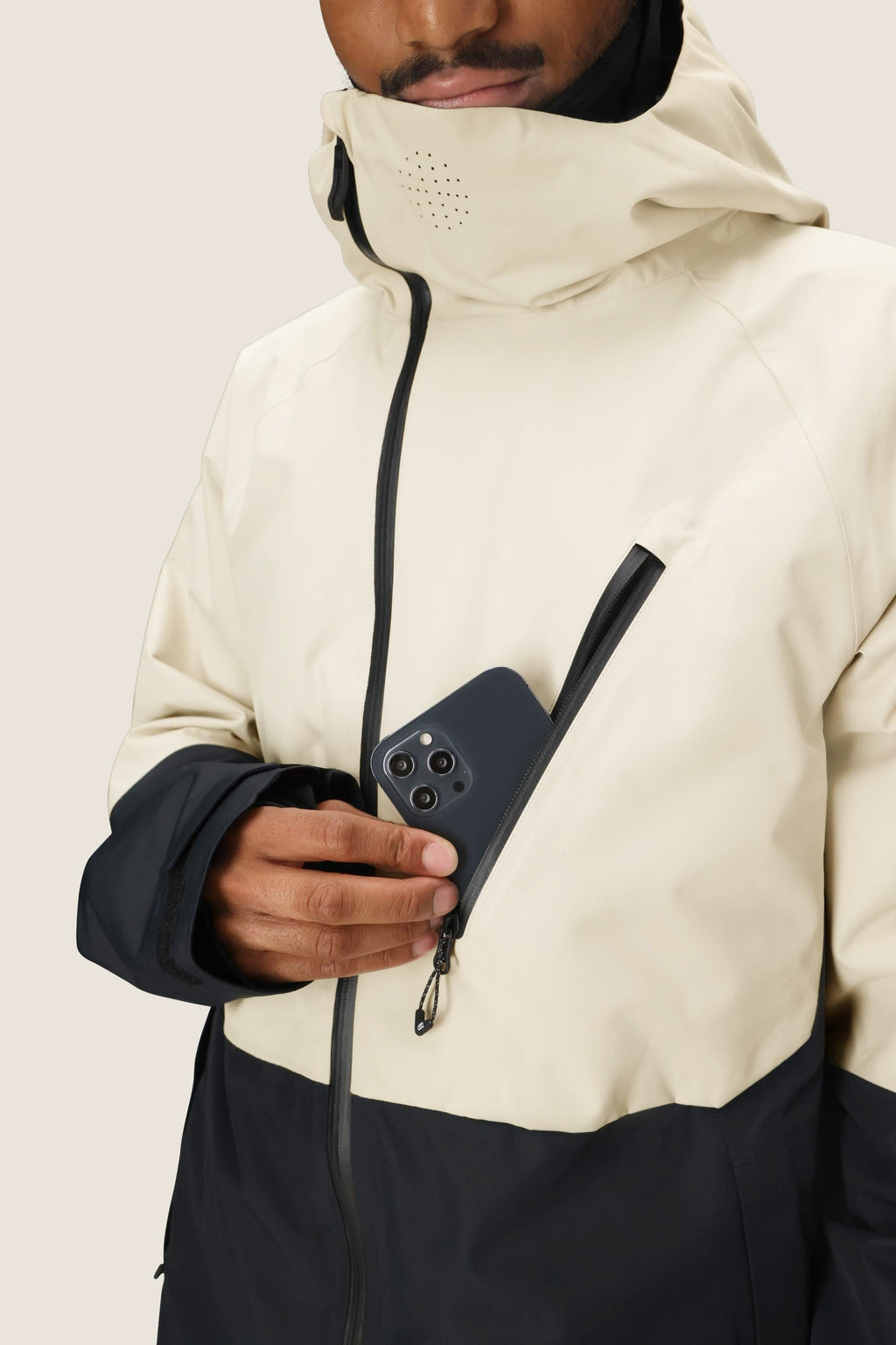 Wearing the 686 Men's Hydra™ Thermagraph® Jacket, featuring a hood and high collar, someone slips a smartphone into the chest pocket. The beige and black 686 jacket offers body-mapped lining for versatile comfort.