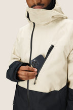 Wearing the 686 Men's Hydra™ Thermagraph® Jacket, featuring a hood and high collar, someone slips a smartphone into the chest pocket. The beige and black 686 jacket offers body-mapped lining for versatile comfort.