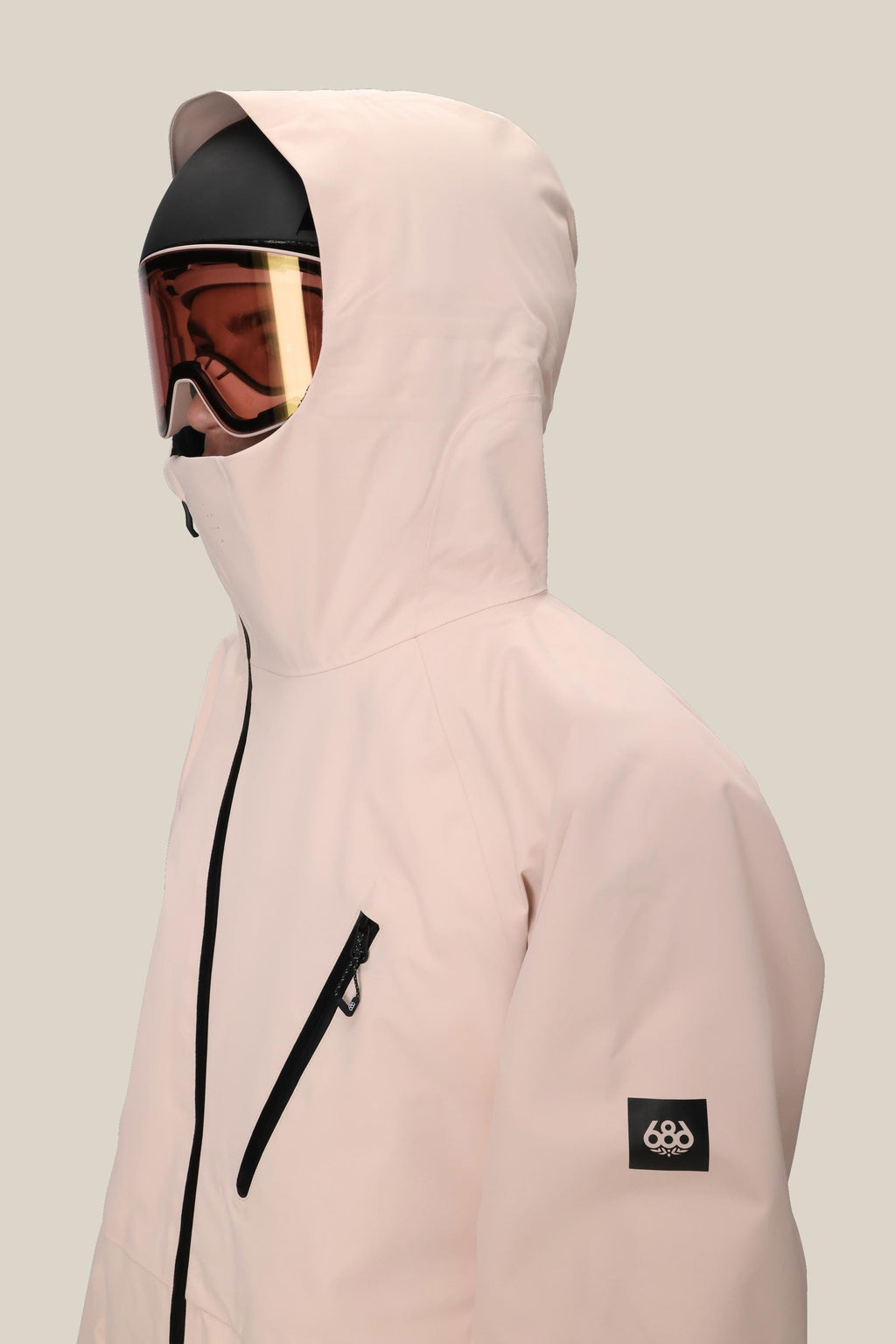 A person wears the 686 Men's Hydra™ Thermagraph® Jacket in light pink with a black zipper, chest pocket, and body-mapped lining. They sport ski goggles and a helmet. The 686 logo appears on the left sleeve against a plain beige background.