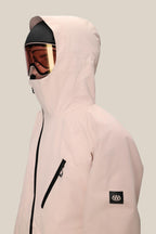 A person wears the 686 Men's Hydra™ Thermagraph® Jacket in light pink with a black zipper, chest pocket, and body-mapped lining. They sport ski goggles and a helmet. The 686 logo appears on the left sleeve against a plain beige background.