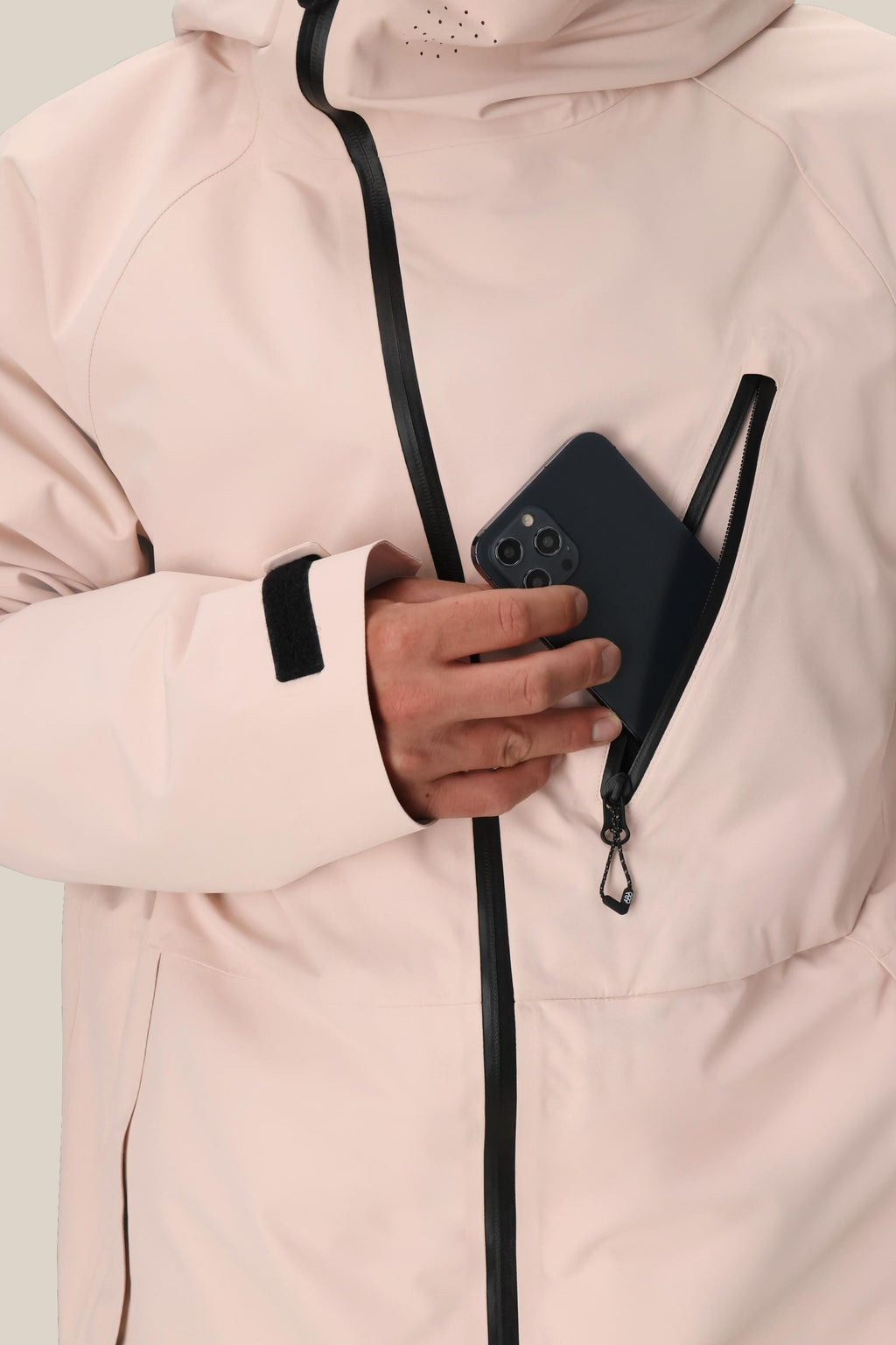 A person wearing the 686 Men's Hydra™ Thermagraph® Jacket in light pink zips a black smartphone into the chest pocket. This best seller from 686 features black zippers, body-mapped lining, and a Velcro sleeve strap.