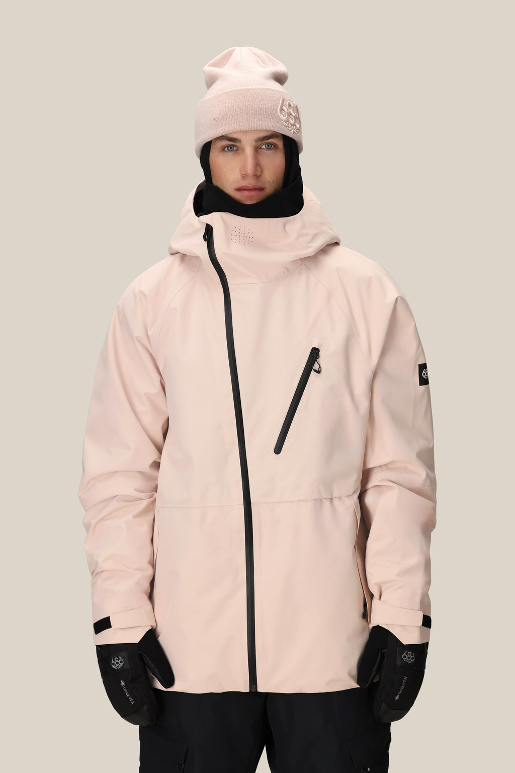 A person wears the 686 Men's Hydra™ Thermagraph® Jacket in light pink with black zippers, a matching pink beanie, black gloves, and black pants while standing against a plain beige background.