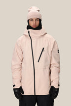 A person wears the 686 Men's Hydra™ Thermagraph® Jacket in light pink with black zippers, a matching pink beanie, black gloves, and black pants while standing against a plain beige background.