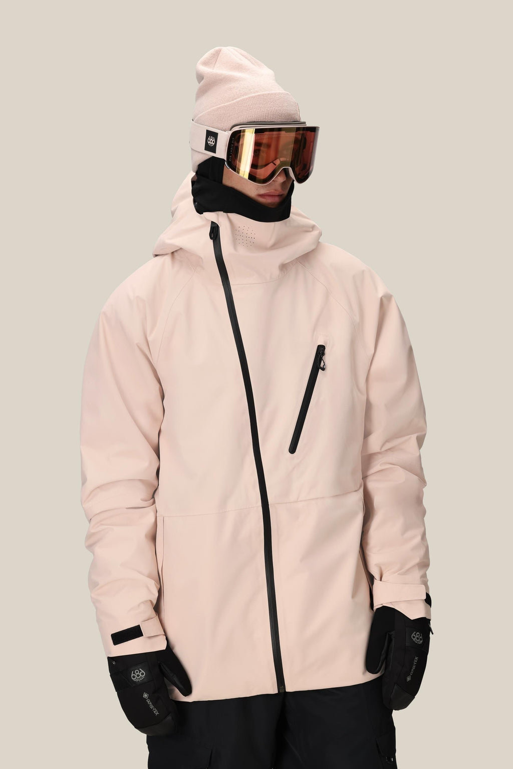 A person wears the 686 Men's Hydra™ Thermagraph® Jacket by 686 with a pink beanie, black gloves, black neck gaiter, and reflective ski goggles, standing against a plain beige background.