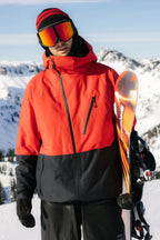 Wearing the 686 Men's Hydra™ Thermagraph® Jacket in red and black, plus a red beanie and reflective ski goggles, a snowboarder stands in snowy mountains holding a snowboard. Brand: 686.