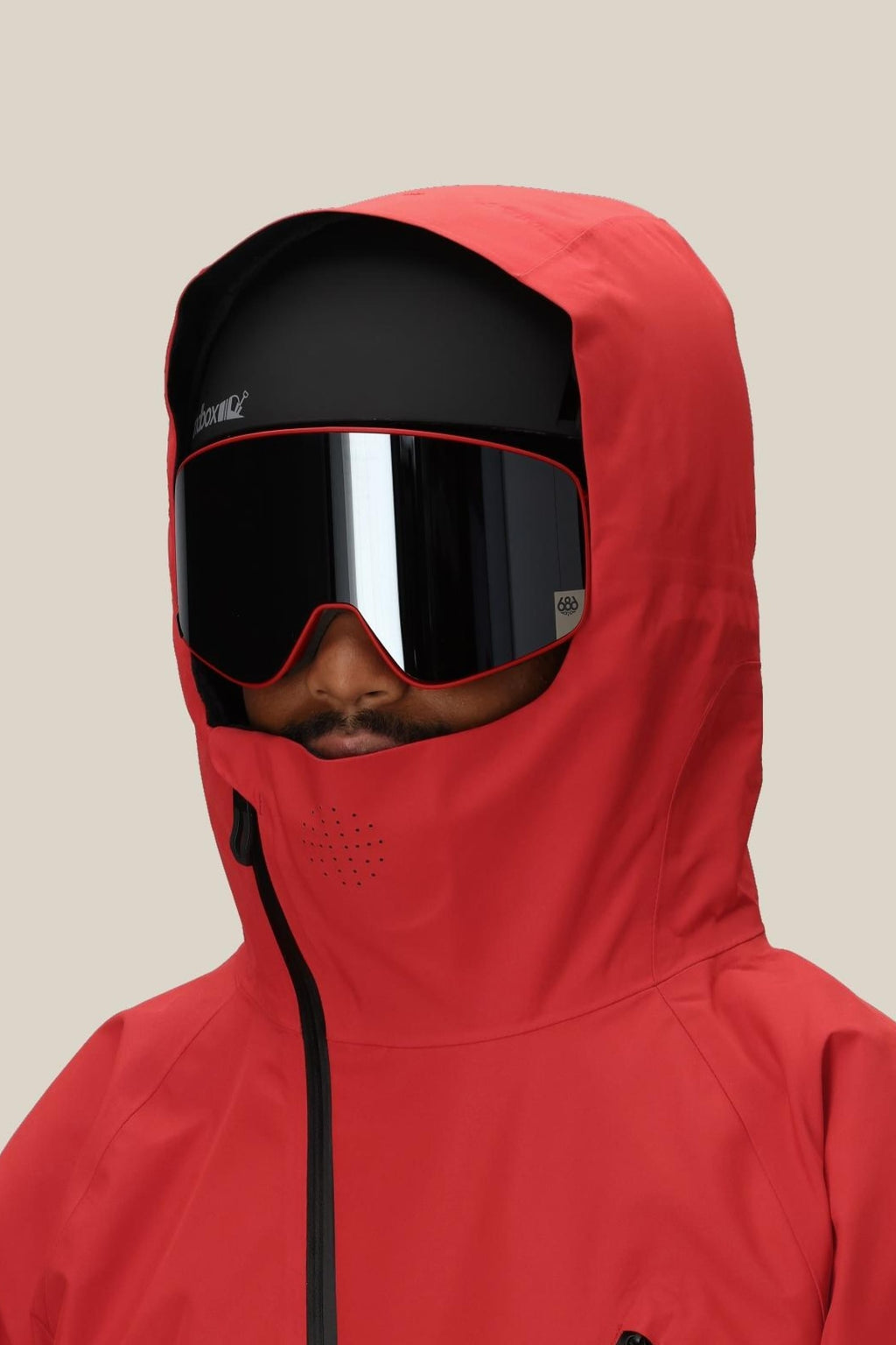A person wearing the 686 Men's Hydra™ Thermagraph® Jacket, a best-selling red hooded jacket by 686, with body-mapped lining and large black ski goggles with red accents stands against a plain beige background.