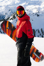 A snowboarder in a best-selling 686 Men's Hydra™ Thermagraph® Jacket with body-mapped lining, black pants, and red goggles stands on snowy mountain terrain, holding a colorful orange and black striped snowboard.