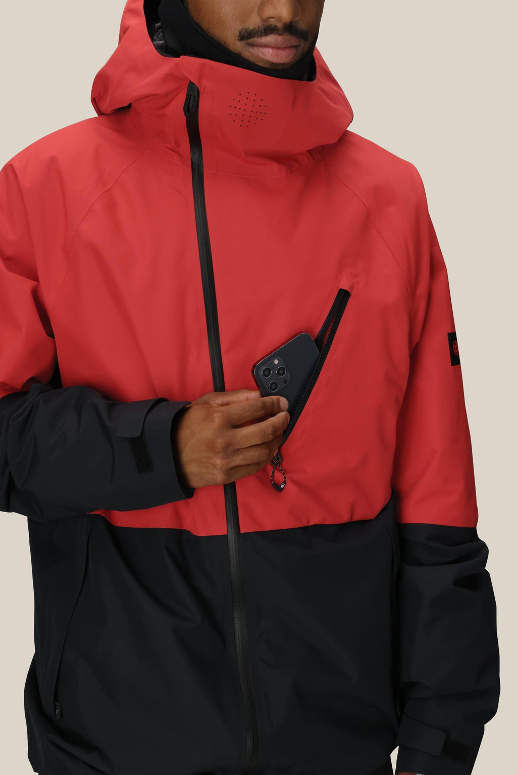 A person wearing the 686 Men's Hydra™ Thermagraph® Jacket with body-mapped lining slips a black phone into the chest pocket, their face partially out of frame against a plain beige background.