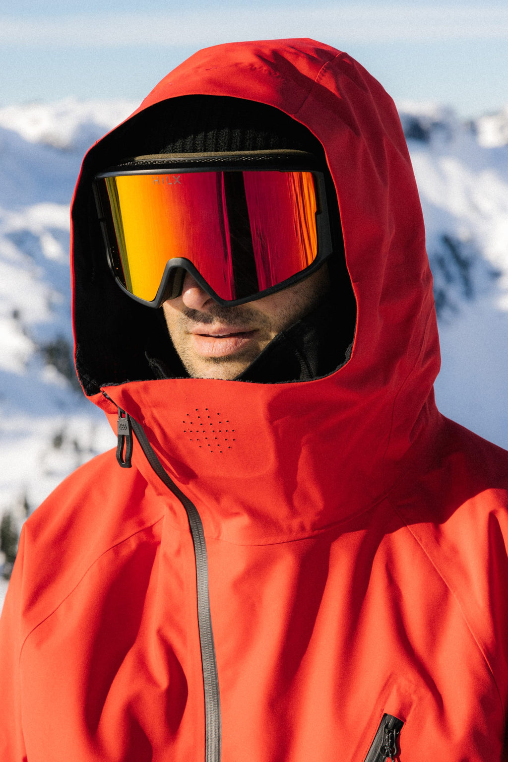 A person wearing a bright red 686 Men's Hydra™ Thermagraph® Jacket, black ski goggles with an orange tint, and a black beanie stands outdoors with snow-covered mountains in the background.