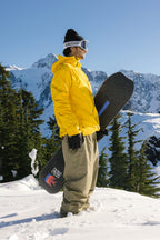 Wearing the 686 Men's Hydra™ Thermagraph® Jacket in yellow, tan pants, and goggles, a snowboarder stands on snowy ground with mountains and evergreens beneath a clear blue sky.