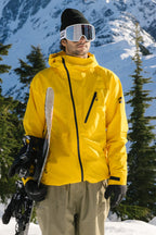 A person wearing the 686 Men's Hydra™ Thermagraph® Jacket by 686 stands in a snowy mountain landscape with ski gear, tan pants, black gloves, and ski goggles, surrounded by pine trees and snow-covered peaks.