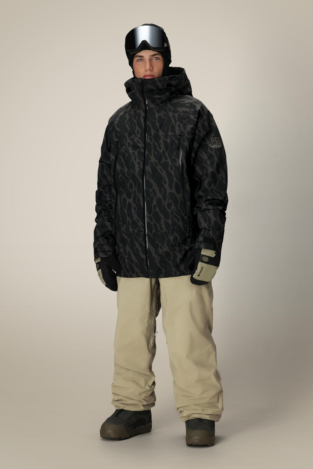 A person wearing a 686 Men's SMARTY® 3-In-1 Rodeo™ 3L Jacket, beige snow pants, black gloves, gray snow boots, and black goggles stands against a plain light background.