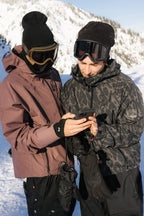 Two people wearing 686 Men's SMARTY® 3-In-1 Rodeo™ 3L Jackets and ski goggles stand together on a snowy mountain, checking a smartphone while the sun shines on snow-covered trees in the background.