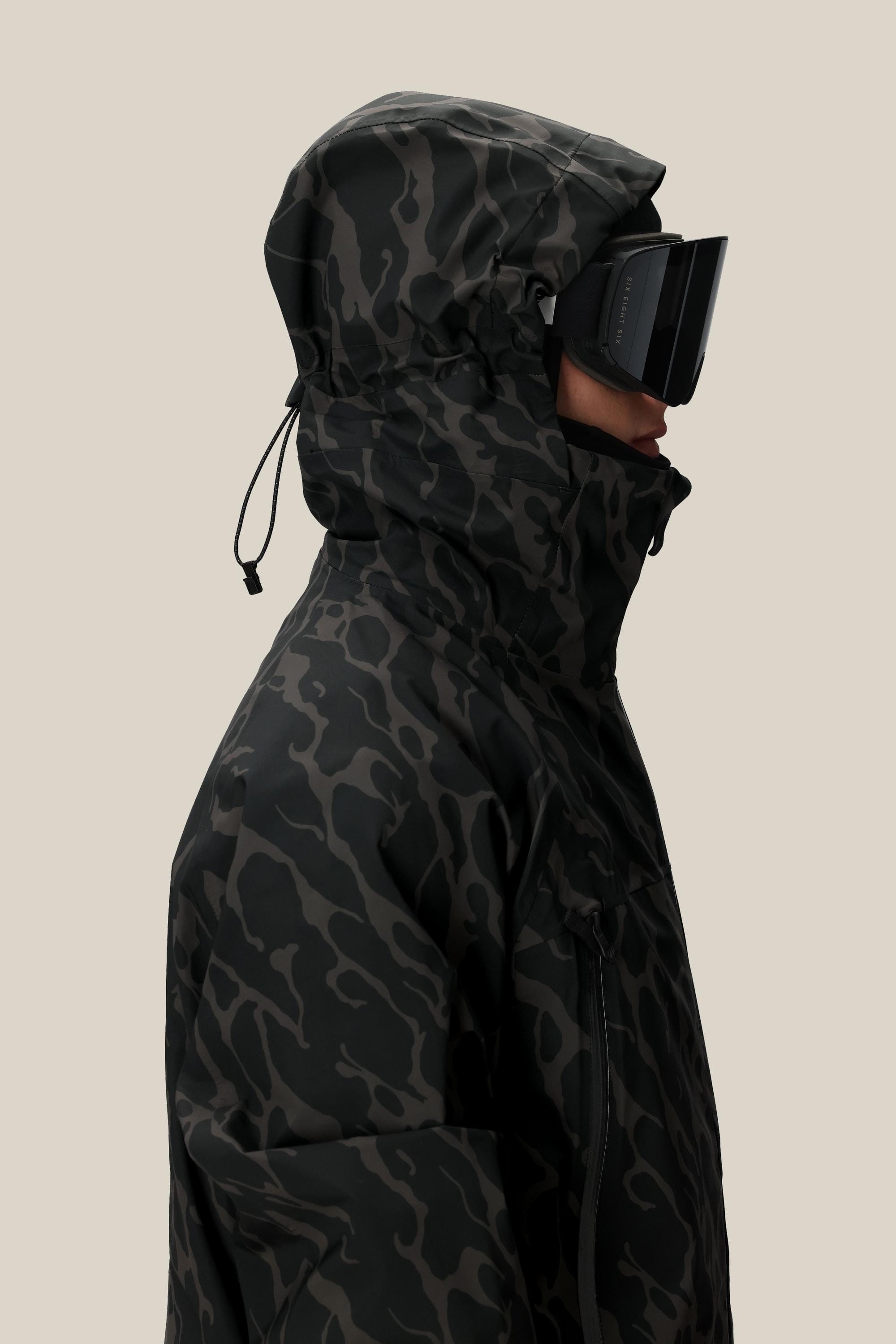 A person in profile wears the 686 Men's SMARTY® 3-In-1 Rodeo™ 3L Jacket with the hood up and black ski goggles, set against a plain beige background.