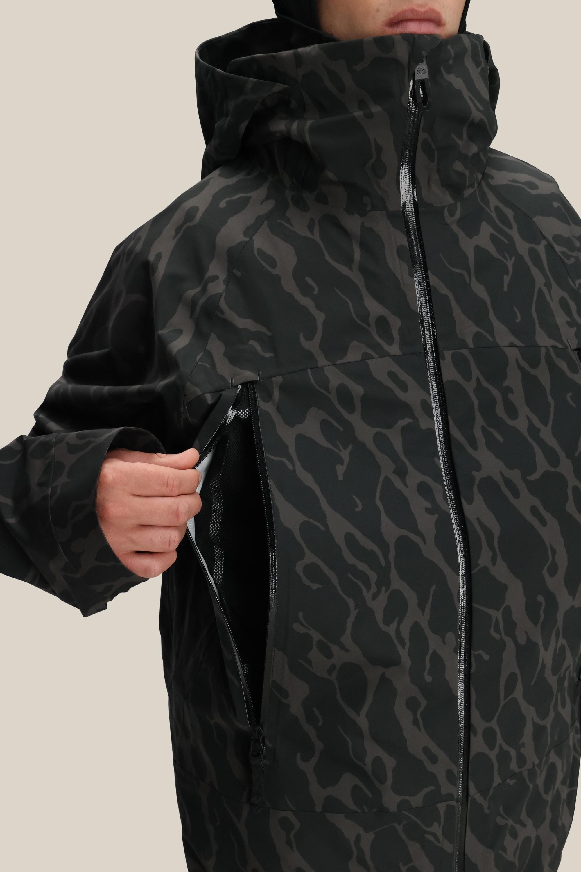 A person in a 686 Men's SMARTY® 3-In-1 Rodeo™ 3L Jacket with dark camo pattern unzips a large side vent pocket on the chest. Their face is partially visible against a plain beige background.