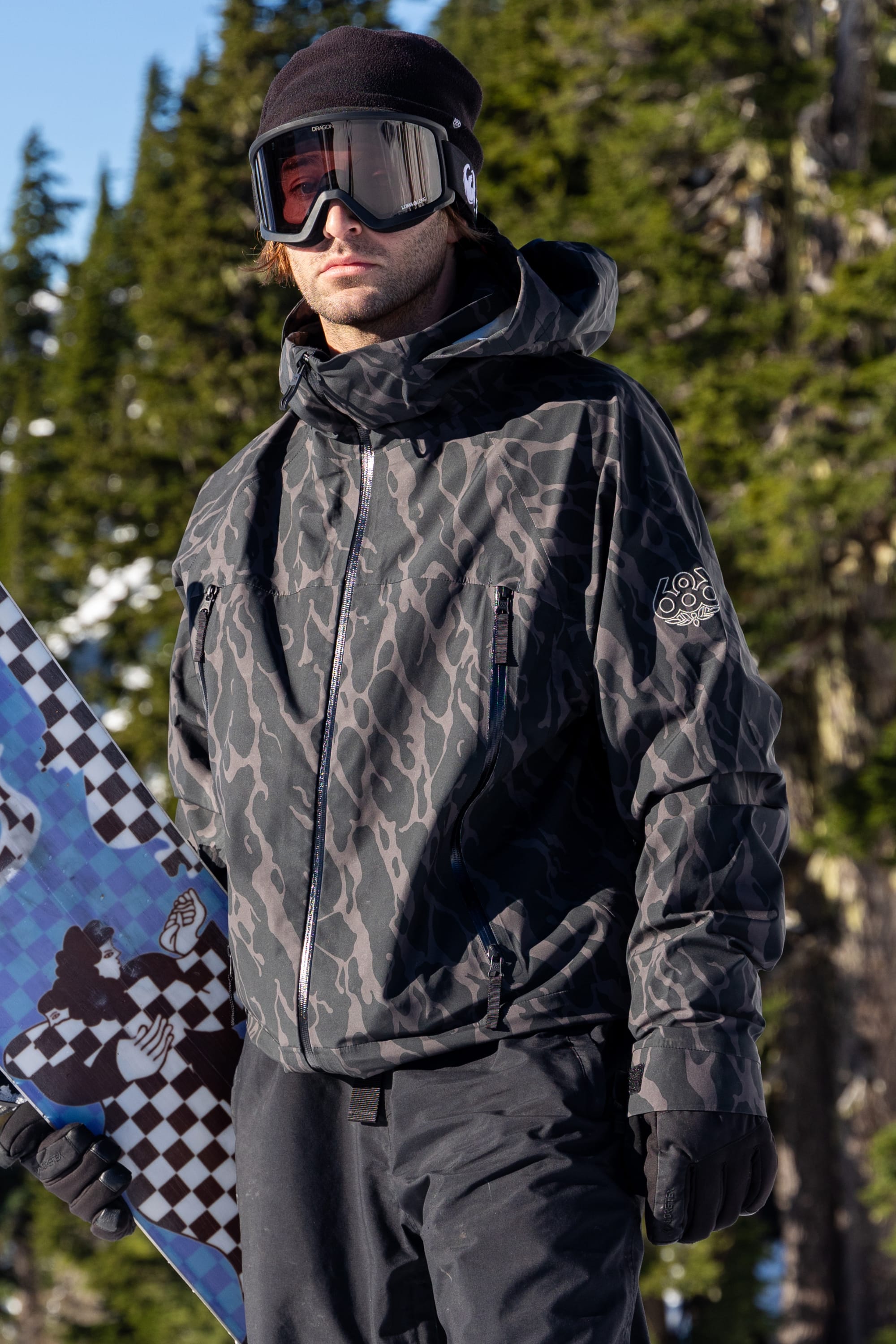 A person wearing the 686 Men's SMARTY® 3-In-1 Rodeo™ 3L Jacket stands outdoors with a snowboard, dressed in black pants and ski goggles. Snow and pine trees are visible in the background.