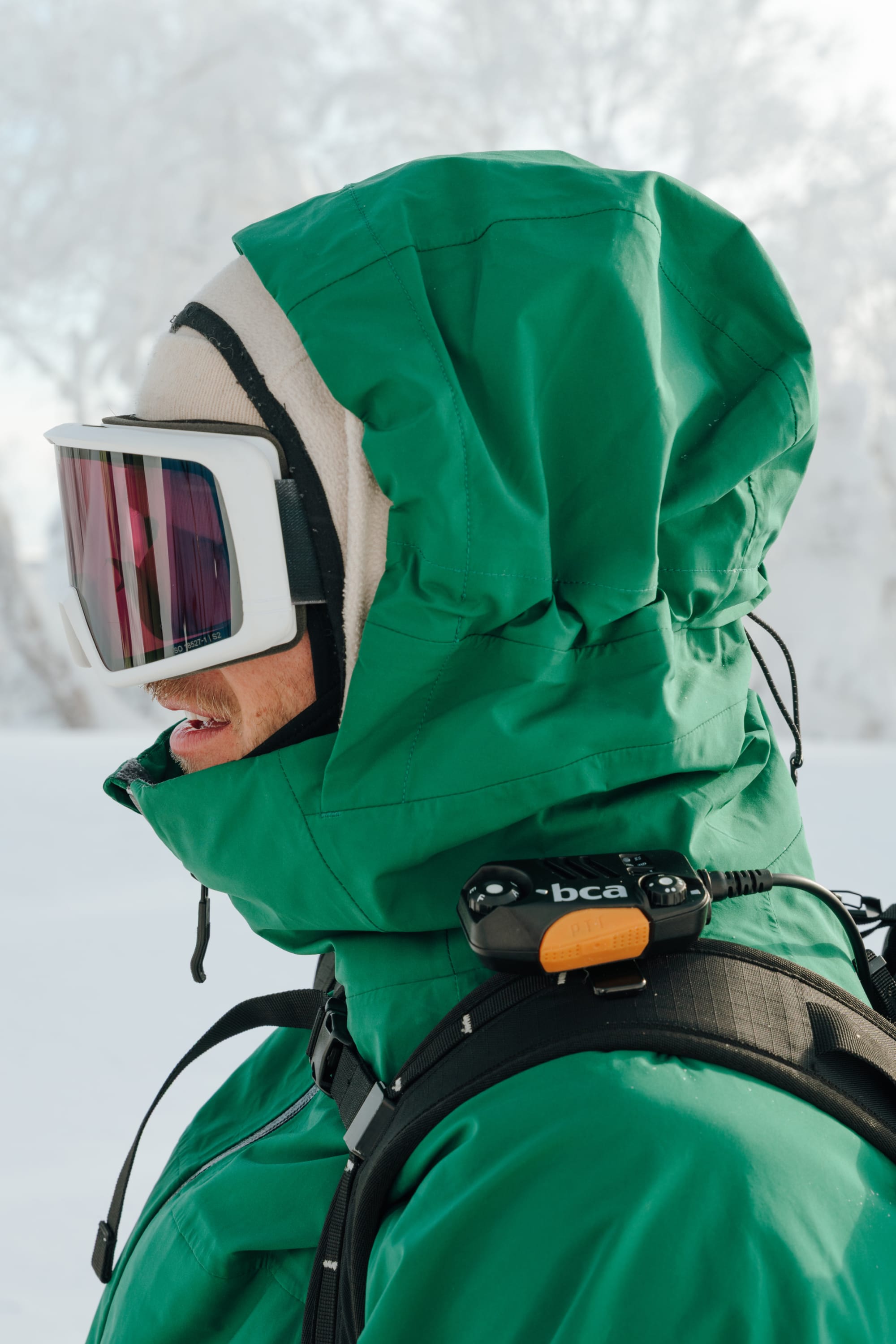 A person wearing a 686 Men's SMARTY® 3-In-1 Rodeo™ 3L Jacket with a green hood, ski goggles, and a beige balaclava stands outdoors in the snow. A black device labeled bca is attached to their backpack strap.