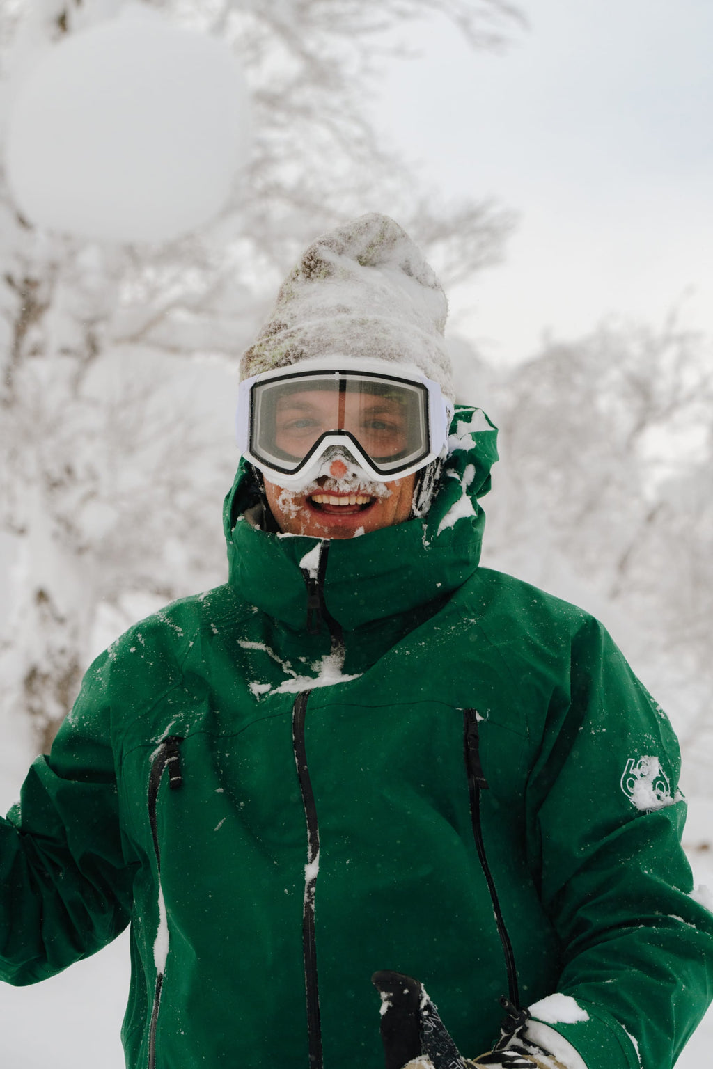 A man wearing a 686 Men's SMARTY® 3-In-1 Rodeo™ 3L Jacket, ski goggles, and a beanie stands smiling outdoors in the snow, with snow on his face and clothes. Snow-covered trees are visible in the background.
