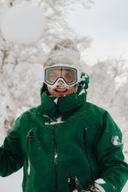 A man wearing a 686 Men's SMARTY® 3-In-1 Rodeo™ 3L Jacket, ski goggles, and a beanie stands smiling outdoors in the snow, with snow on his face and clothes. Snow-covered trees are visible in the background.