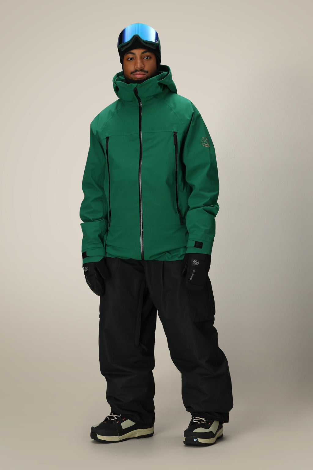 A person wearing a green 686 Men's SMARTY® 3-In-1 Rodeo™ 3L Jacket, black snow pants, black gloves, black-and-white snow boots, and a blue ski visor stands against a plain light background.