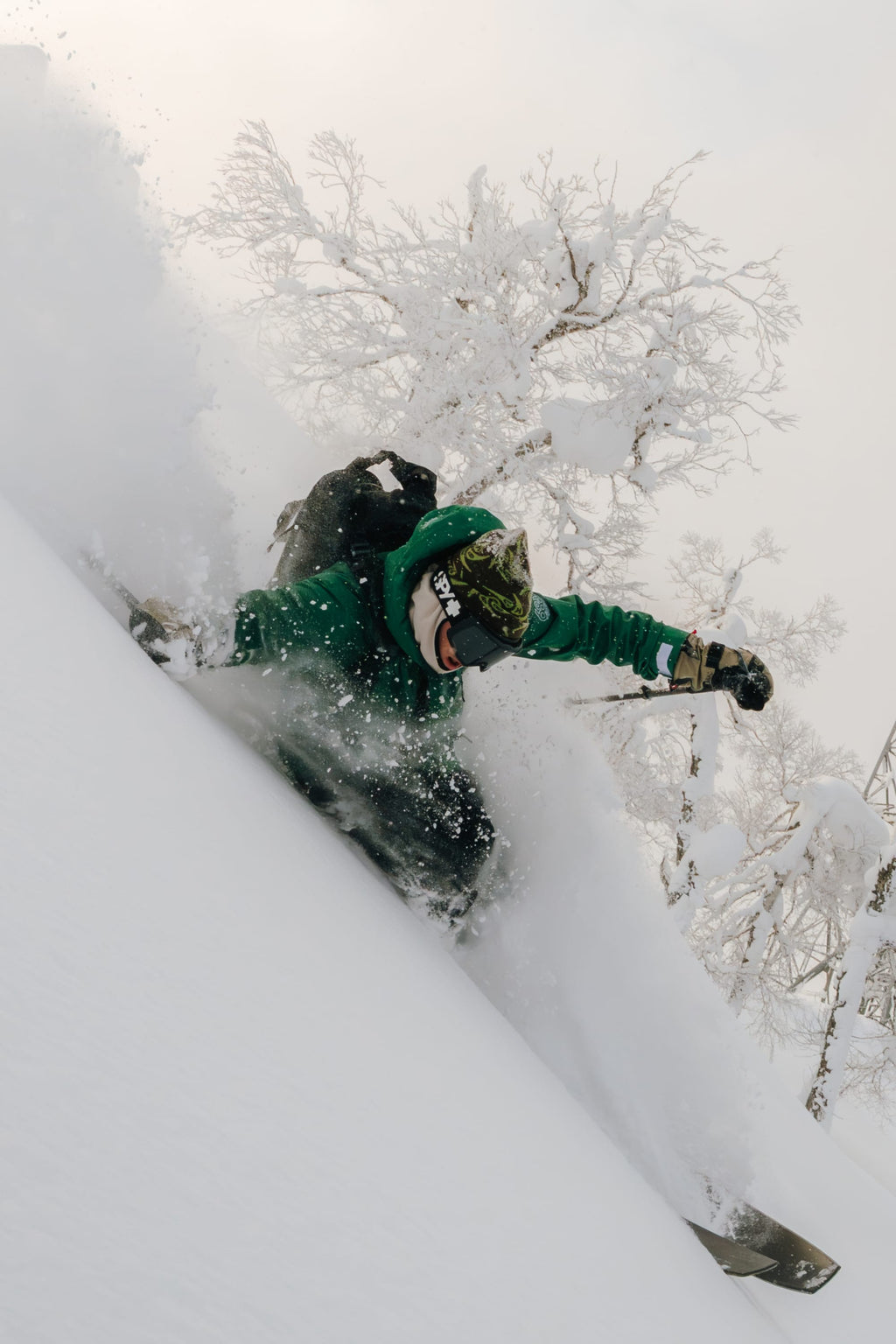 A snowboarder wearing the 686 Men's SMARTY® 3-In-1 Rodeo™ 3L Jacket and helmet carves through deep powder on a snowy slope, with snow-covered trees in the background.