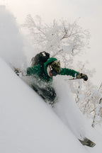 A snowboarder wearing the 686 Men's SMARTY® 3-In-1 Rodeo™ 3L Jacket and helmet carves through deep powder on a snowy slope, with snow-covered trees in the background.