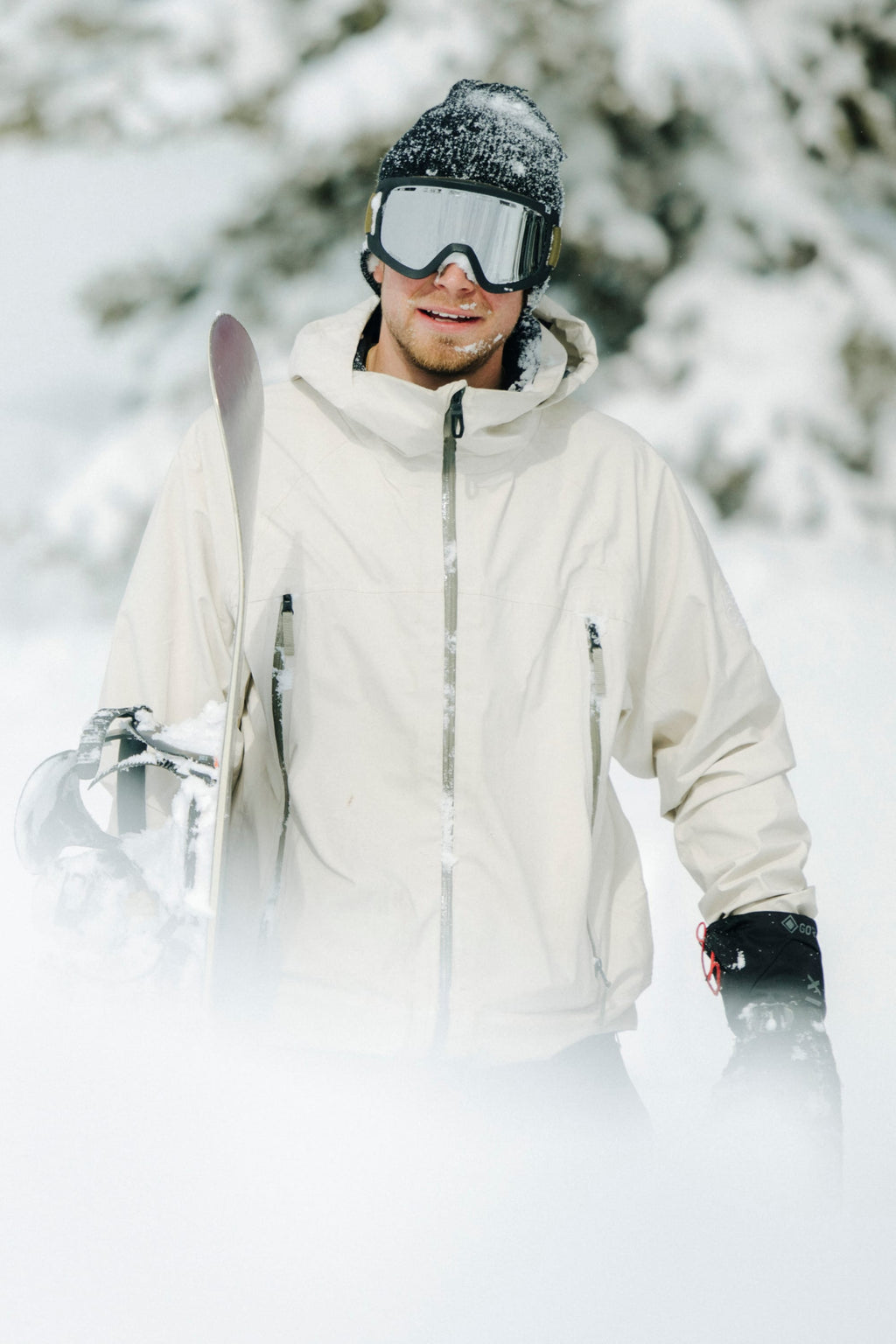 Wearing the 686 Men's SMARTY® 3-In-1 Rodeo™ 3L Jacket, a person stands in a snowy landscape with skis in hand. Snow blankets the ground and trees while they complete their look with black gloves and goggles.