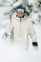 Wearing the 686 Men's SMARTY® 3-In-1 Rodeo™ 3L Jacket, a person stands in a snowy landscape with skis in hand. Snow blankets the ground and trees while they complete their look with black gloves and goggles.
