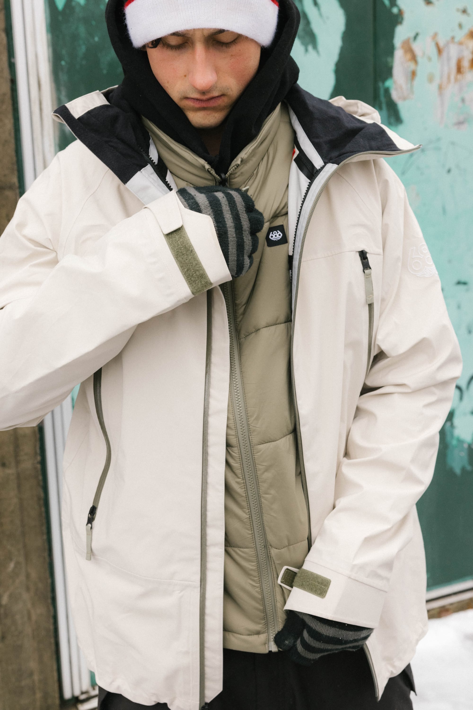 A person wearing the 686 Men's SMARTY® 3-In-1 Rodeo™ 3L Jacket in white, an olive green puffer vest, black and white striped gloves, and a white beanie stands by a weathered green wall, looking down while zipping up their jacket.