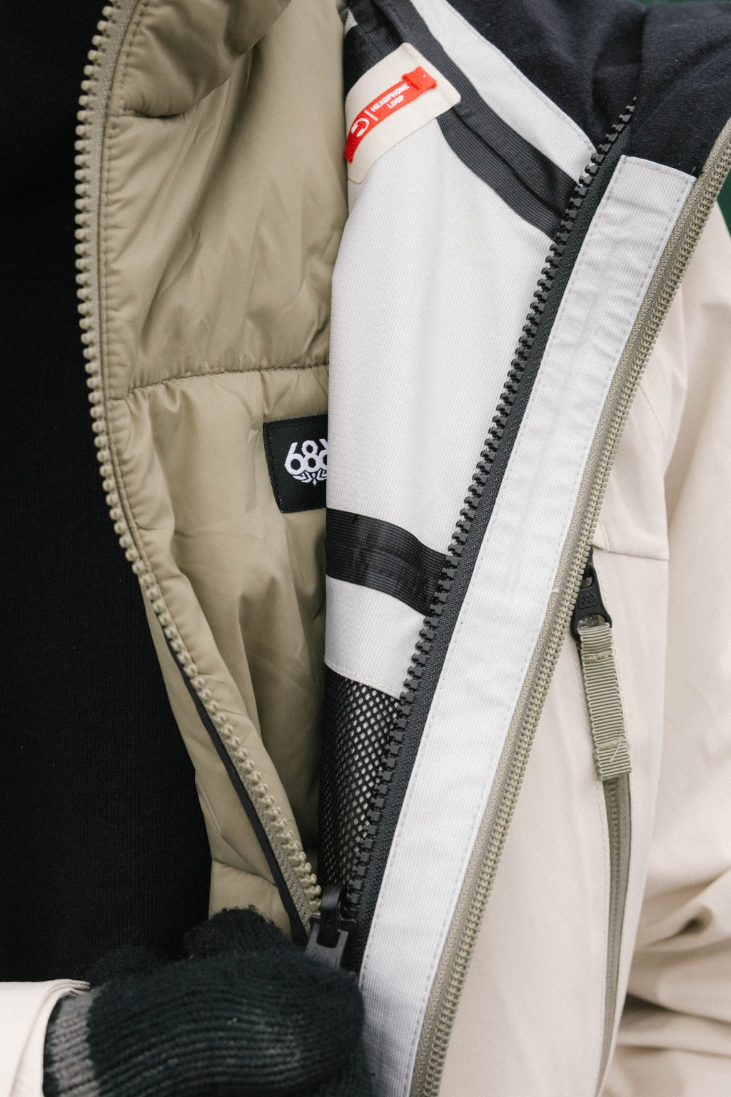 A close-up of a person wearing the 686 Men's SMARTY® 3-In-1 Rodeo™ 3L Jacket in beige and white, partially unzipped to show the interior lining and 686 label. The person also wears a black sweater and black gloves.