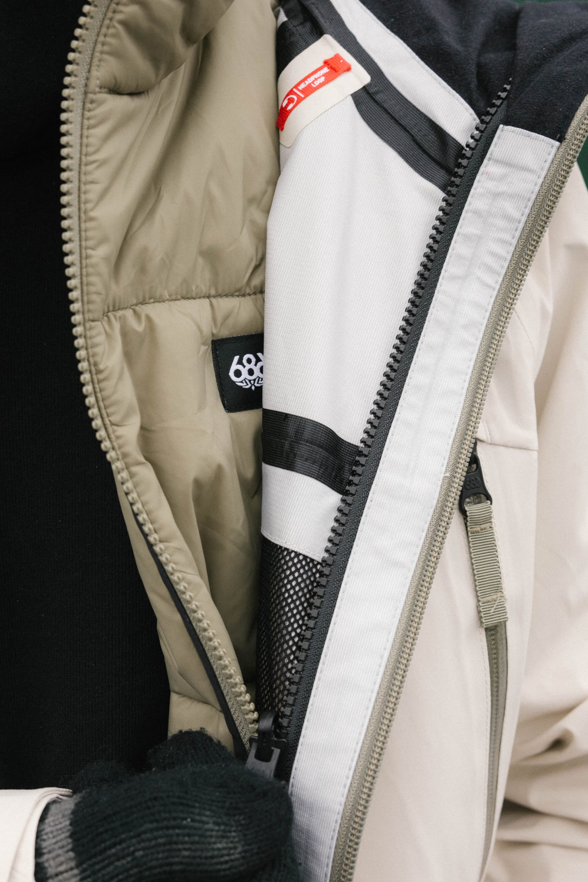 A close-up of a person wearing the 686 Men's SMARTY® 3-In-1 Rodeo™ 3L Jacket in beige and white, partially unzipped to show the interior lining and 686 label. The person also wears a black sweater and black gloves.