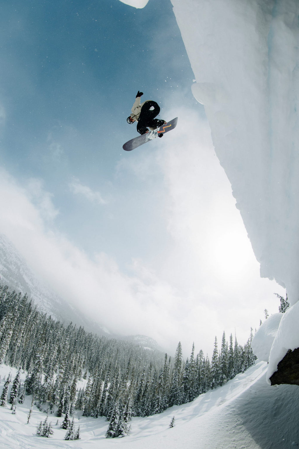 A snowboarder in the 686 Men's SMARTY® 3-In-1 Rodeo™ 3L Jacket launches off a snowy cliff, soaring mid-air with snow-covered trees and mountains behind him on a bright, cloudy day.