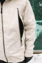 A man in a light beige 686 Men's SMARTY® 3-In-1 Rodeo™ 3L Jacket, featuring a large vertical zippered pocket, stands in front of a green and white textured wall with only his torso and one arm visible.