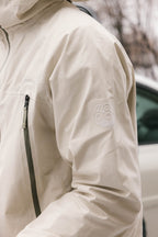A close-up of a person wearing the 686 Men's SMARTY® 3-In-1 Rodeo™ 3L Jacket, featuring a zippered chest pocket and a white 686 logo on the sleeve. In the background, there's a blurred car and trees.