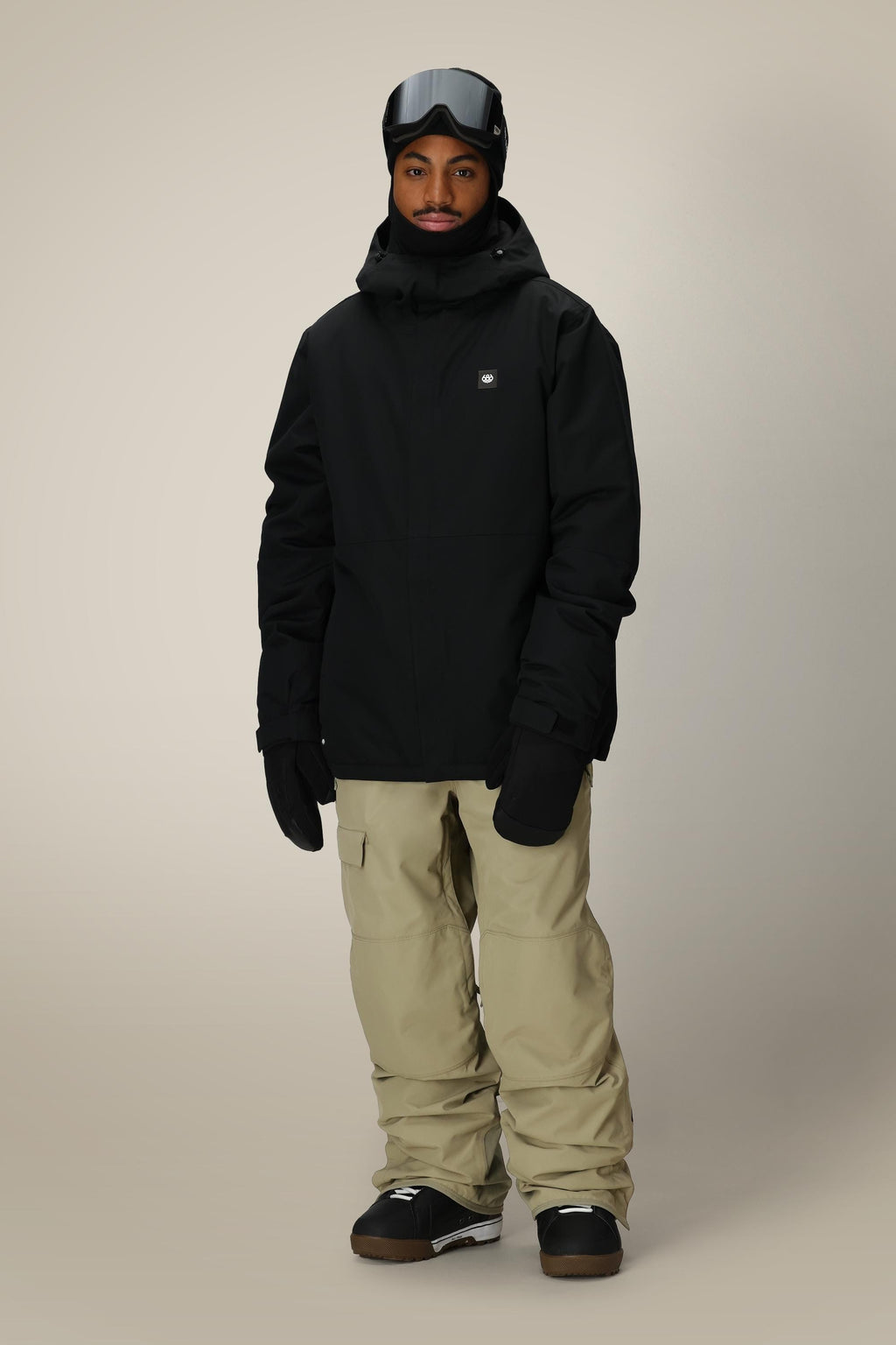 A person wearing the 686 Men's Foundation™ Insulated Jacket, beige snow pants, black gloves, black snow boots, and ski goggles stands facing forward against a plain light background.