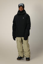 A person wearing the 686 Men's Foundation™ Insulated Jacket, beige snow pants, black gloves, black snow boots, and ski goggles stands facing forward against a plain light background.