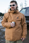 A man wearing a 686 Men's Foundation™ Insulated Jacket and sunglasses stands outdoors holding a can. Snow covers the ground, with a dark vehicle and trees visible in the background.