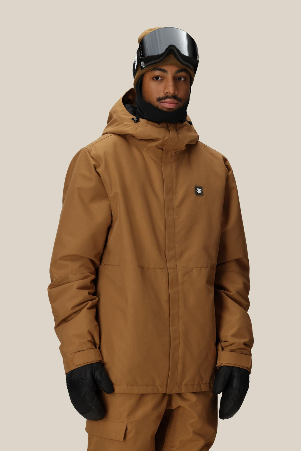 A man wearing a 686 Men's Foundation™ Insulated Jacket in brown, matching pants, black gloves, a black neck warmer, and ski goggles stands against a plain beige background.
