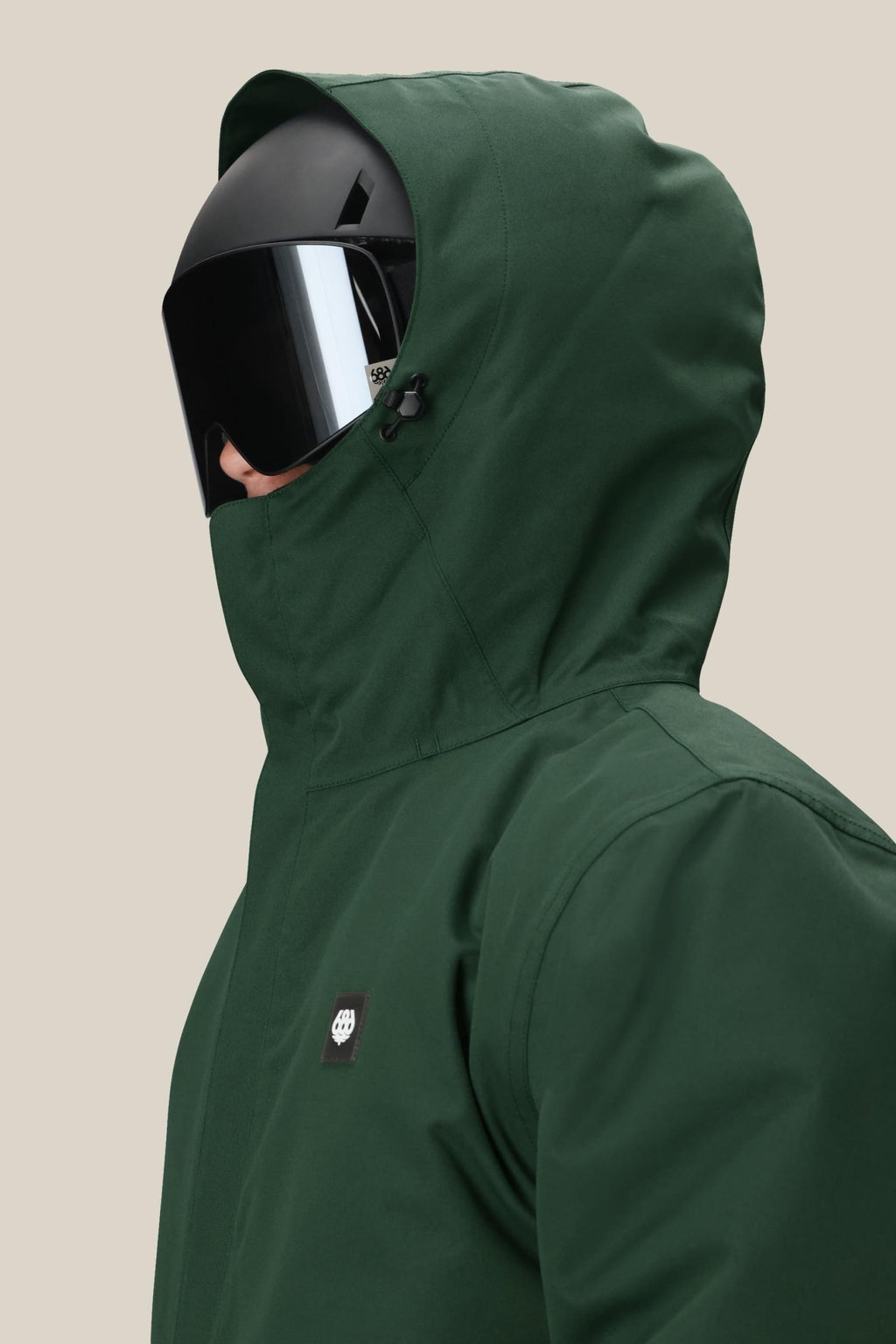 A person wearing the 686 Men's Foundation™ Insulated Jacket in dark green with a hood and a large black visor helmet, facing left against a plain beige background.