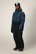 A person stands facing forward, wearing the 686 Men's Foundation™ Insulated Jacket by 686 in a blue and black pattern, along with black snow pants, gloves, snow boots, and ski goggles on their forehead against a plain light background.